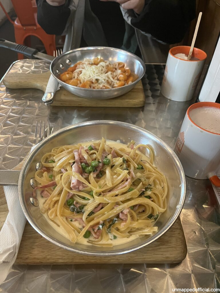 Ham and peas fettucine alfredo served in a pan and a mug of hot chocolate to the side. On the other side of the table, tomato and cheese gnocchi.