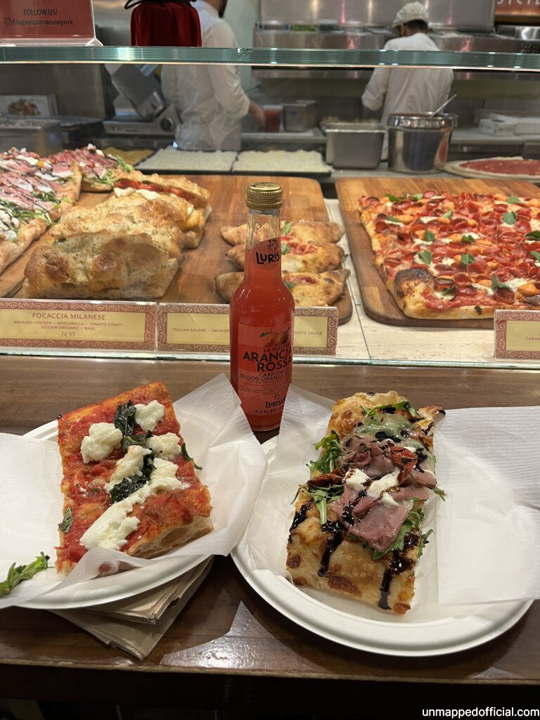 two slices of pizzas with a blood orange soda on a counter with more pizza in the background behind the glass